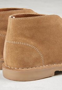 Ruskindsankelstøvler i tan med et afrundet design, kontrasterende hvide syninger og en solid gummisål. Glat tekstur og buede kanter.