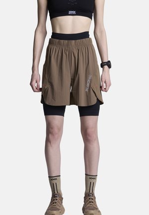 Atleet draagt een zwarte sportbeha, bruine gelaagde korte broek met "X BIONIC"-logo, beige sokken, wandelschoenen en een zwart polshorloge aan de linkerarm.