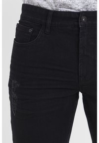 Musta kitsaste lõikega teksapüksid, millel on kulunud piirkond ja standardne viie taskuga disain. Vöökohal nööpidega kinnitamine.