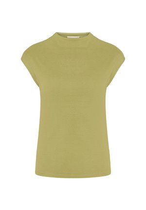 Vrouw met steil donker haar die een mouwloos olijfgroen topje draagt, wijd uitlopende lichtgroene broek en zwarte sandalen met hakken tegen een effen achtergrond.