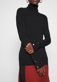 Femme portant un pull à col roulé noir avec des boutons dorés aux poignets et une jupe plissée rouge et noire, les bras croisés.