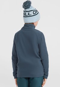Blå fleecepullover med hög krage, med långa ärmar. Kombinerad med en ljusblå stickad mössa med en pom-pom på toppen.