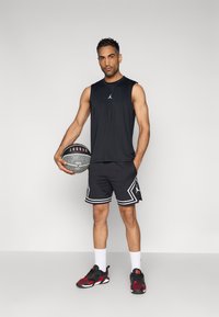 Μαύρο αμάνικο αθλητικό τοπ με λογότυπο, μαύρο σορτς με λευκό περίγραμμα, λευκές κάλτσες και κόκκινα-μαύρα αθλητικά παπούτσια. Κρατάει μια μπάλα του μπάσκετ.