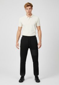 Kremowa męska koszulka polo z trzema guzikami, do tego czarne spodnie dresowe z kieszenią na zamek i teksturowanym panelem. Całość dopełniają czarne sneakersy.