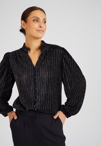 Blusa nera con righe verticali e una leggera lucentezza, dotata di un colletto alto e maniche lunghe a sbuffo. Chiusura frontale con bottoni.