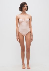 Kobieta stojąca boso, ubrana w jasnoróżowy jednoczęściowy kostium kąpielowy z cienkimi ramiączkami i miseczkami na fiszbinach, na tle jednolitego, jasnego tła.