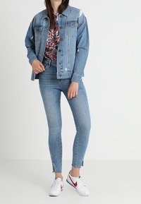 Giacca di jeans blu chiaro con dettagli strappati, abbinata a una camicia floreale e jeans skinny con spacchi laterali. Calzature: sneakers bianche.