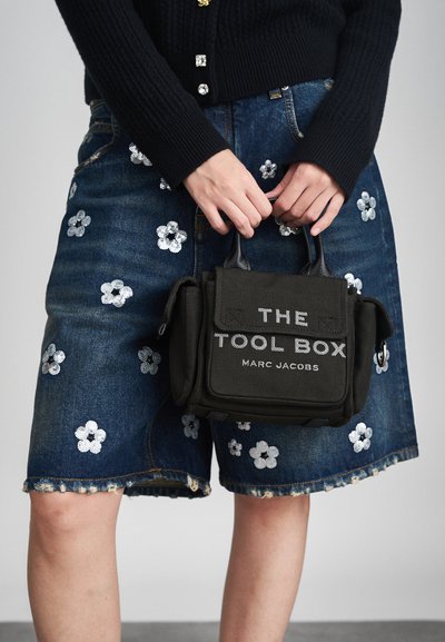 Sorte lærredshåndtaske med "THE TOOL BOX" og "MARC JACOBS" trykt i hvidt. Båret med denimshorts med hvide blomsterdekorationer.