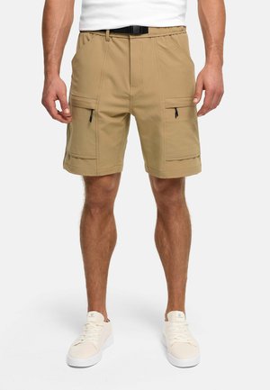 Mężczyzna w jasnobrązowych szortach cargo z przednimi kieszeniami na zamek błyskawiczny i czarnym paskiem, w połączeniu z białymi trampkami i białą koszulką.