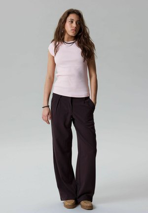 Giovane donna con capelli lunghi castani che indossa una maglia aderente rosa chiaro, pantaloni neri a gamba larga, scarpe color cuoio e collana e braccialetto neri con perline.