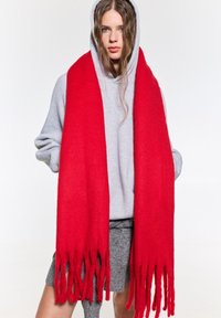 Rode wollen sjaal met franjes aan de uiteinden, gedragen over een grijze hoodie, textuurvol en dik voor warmte.