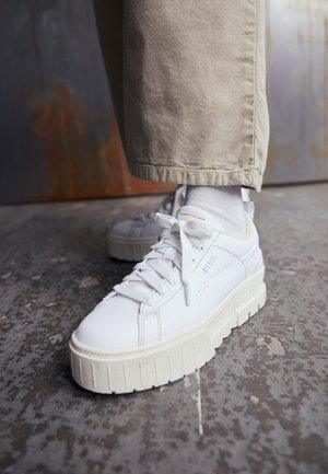 Białe sneakersy Puma na platformie, zasznurowane, noszone z białymi skarpetkami i beżowymi spodniami, stojące na teksturowanej szarej podłodze.