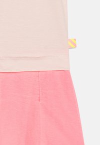 Jasnoróżowa bluzka z krótkimi rękawami w zestawieniu z intensywnie różową spódnicą. Posiada wszytą metkę z ukośnymi żółtymi i różowymi paskami.