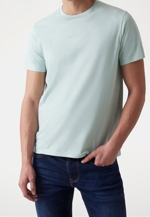 Jasnoniebieska bawełniana koszulka z okrągłym dekoltem i krótkimi rękawami, z ton w ton logo "GUESS" na piersi. Noszona z ciemnymi dżinsami.