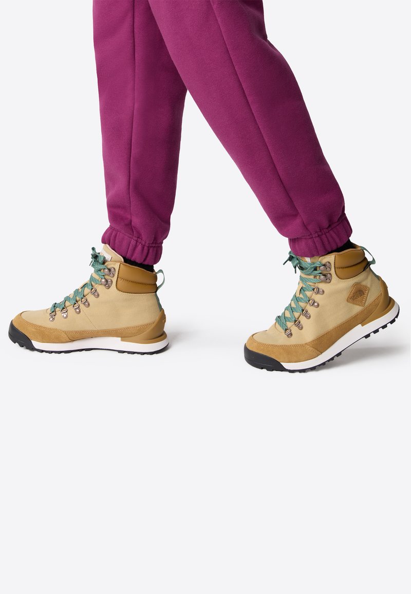 Beige Wanderschuhe mit grünen Schnürsenkeln, Obermaterial aus Wildleder und Mesh, Gummisohle und Metallösen, kombiniert mit lila Fleece-Jogginghosen.