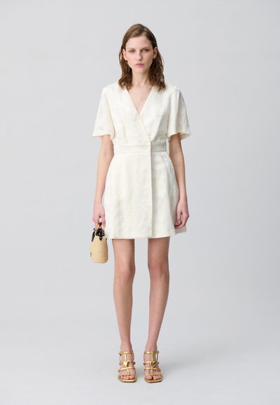 Robe crème à manches courtes avec un design croisé, taille cintrée et tissu texturé. Associée à des sandales à lanières marron et à un petit sac tissé.