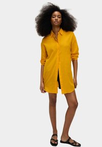 Camicia a maniche lunghe, con bottoni, in lino giallo brillante, con una vestibilità rilassata e colletto. Indossata sopra dei pantaloni corti neri, con sandali piatti.