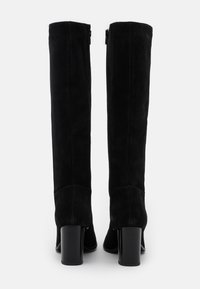 Un par de botas de ante negras hasta la rodilla con tacones cuadrados mostradas desde atrás sobre un fondo blanco.