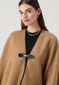 Kamelfarvet uld poncho med sorte læderdetaljer og en guldspænde lukning. Har en designet halsudskæring og en glat tekstur.