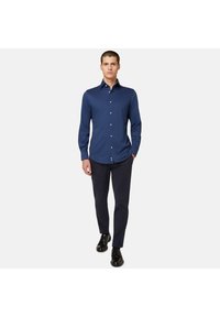 Mornarsko modra srajca z dolgimi rokavi z ovratnikom na gumb, s kontrastnimi belimi gumbi, v kombinaciji s temnimi hlačami in črnimi čevlji.