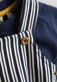Pruhovaný tmavomodro-biely materiál s dreveným gombíkom na zapínanie, doplnený žltým vnútorným golierom. Mäkká textura, vhodná pre deti.