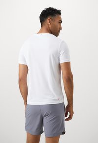 Weißes, kurzärmliges Sportshirt aus strukturiertem Stoff mit einem rosa Akzent am Saum, kombiniert mit grauen Shorts. Rückansicht gezeigt.