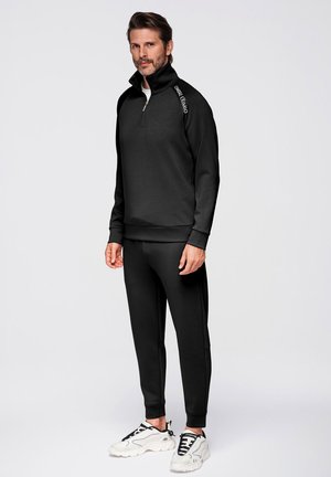 Mężczyzna stojący, ubrany w czarny pulower z suwakiem do ćwiartki z napisem "OMBRE EXXIMO" na ramieniu, czarne spodnie i białe trampki na szarym tle.