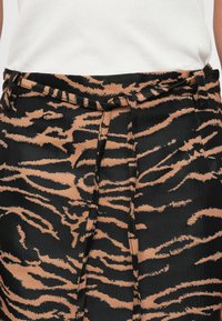 Hoge taille broek in een zwarte stof met een tan kleurige tijgerprint, voorzien van een ceintuurlus en een geplooide voorkant. Zachte textuur.