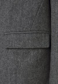 Närbild av grått herringbone-mönstrat kostymtyg med en horisontell fickdetalj.