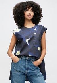 Tengerészkék ujjatlan felső absztrakt sárga és fehér mintákkal, kerek nyakkivágással és mellzsebbel; világoskék farmerral párosítva.