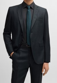 Completo scuro con motivo a fantasia e design aderente, abbinato a una cravatta verde acqua tinta unita e una camicia nera. Dettagli sartoriali e silhouette strutturata.