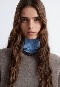 Brauner Wollpullover mit hohem, geripptem, hellblauem Kragen. Eine dunkle Perlenkette liegt am Kragenansatz. Langes, welliges Haar umrahmt das Gesicht.