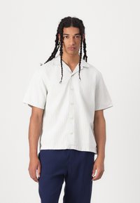 Chemise à manches courtes, de couleur claire et à rayures, avec un devant boutonné, une coupe décontractée et une texture de tissu douce. Portée avec un pantalon bleu foncé.