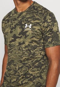 Camiseta verde con camuflaje que presenta un patrón en píxeles negros, cuello redondo y logo blanco de "Under Armour" en el pecho. Manga corta.