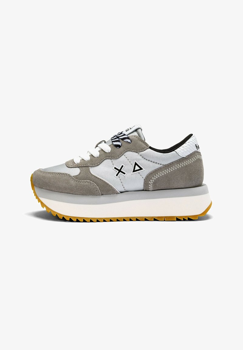 Zapatilla gris y blanca con materiales de gamuza y tela, que presenta detalles en negro, una suela texturizada y diseños geométricos en los laterales.