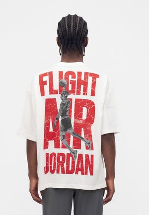 Person mit geflochtenem Haar, die ein weißes T-Shirt trägt, auf dem ein schwarz-weißer Basketballspieler zu sehen ist, der mit einem Ball über großen roten Text "Flight Air Jordan" springt.