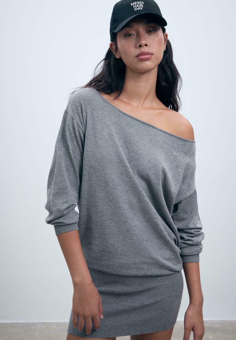 Robe sweat gris avec design dénudé d'épaule, manches longues, texture côtelée à l'ourlet. Casquette noire avec le texte « JOUR DE CHEVEUX EN DÉSORDRE ».