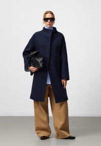 Manteau en laine navy avec un col haut et des boutons sur le devant. Associé à une chemise bleu clair, un pantalon large beige et un sac à main noir.