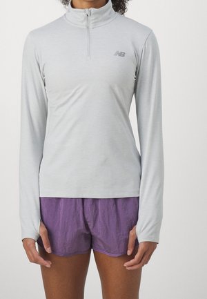 Person, der einen hellgrauen, langärmligen Sportpullover mit Viertelreißverschluss und lila Laufshorts trägt, vor einem einfarbigen Hintergrund steht.