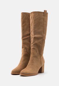 Bottes hautes en daim beige avec un bout pointu et un talon bloc. Elles disposent d'une fermeture éclair latérale et d'une boucle pour un enfilage facile. Surface à texture lisse.