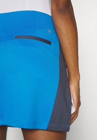 Modrá a šedá sportovní sukně s texturovanou tkaninou, přiléhavým střihem a malou skrytou kapsou na boku s kontrastním lemem.