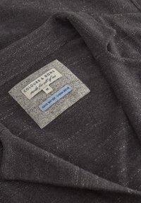Tessuto grigio scuro con una leggera texture, con un'etichetta che recita "COLOURS & SONS" e ulteriori etichette riguardanti taglia e istruzioni per la cura.