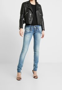 Svart lädermotorcykeljacka med ribbade axeldetaljer, tillsammans med ljusblå tajta jeans. Modellen står i svarta klackskor.