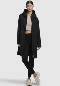 Manteau imperméable noir avec capuche, fermeture à boutons sur le devant et deux poches latérales. Associé à des leggings noirs et des baskets de couleur neutre.
