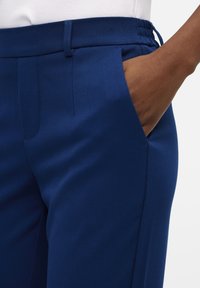 Marineblauwe stretchbroek met een gladde textuur, voorzien van een ontspannen tailleband en zijzakken. Modieuze snit met subtiele plooien.