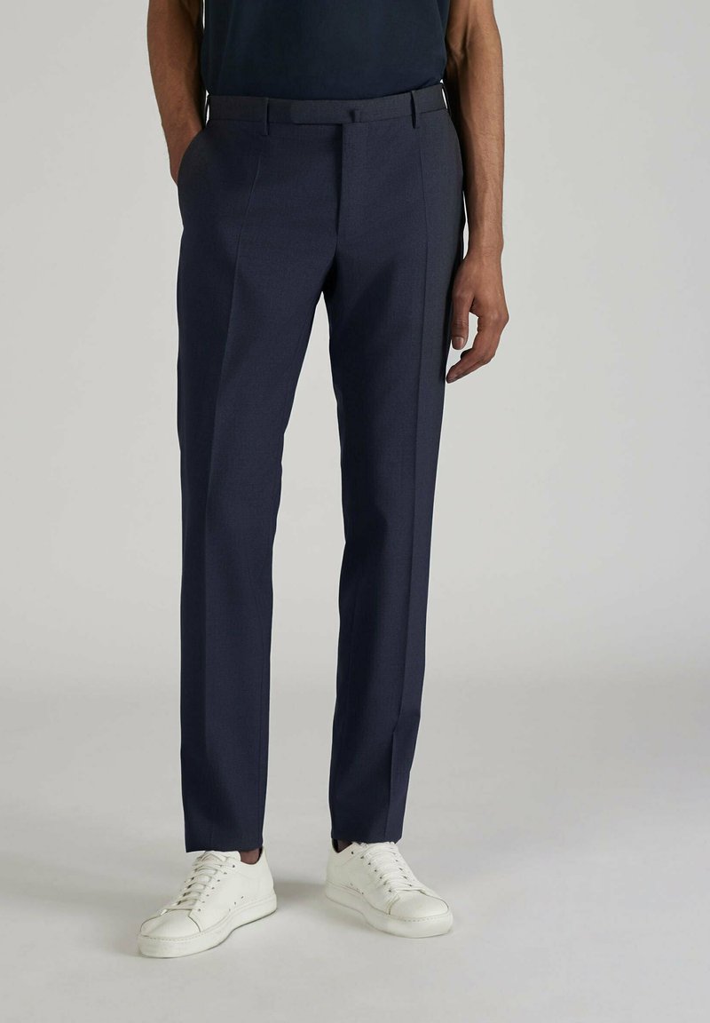 Pantalons de costume bleu marine en tissu lisse avec une coupe ajustée, dotés d'un design à plat et de poches sur les côtés, associés à des baskets blanches.