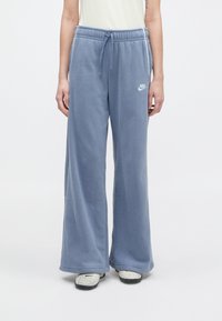 Άτομο ντυμένο με χαλαρούς ανοιχτό γαλάζιους φλις παντελόνες Nike και λευκά αθλητικά παπούτσια μπροστά σε απλό λευκό φόντο.