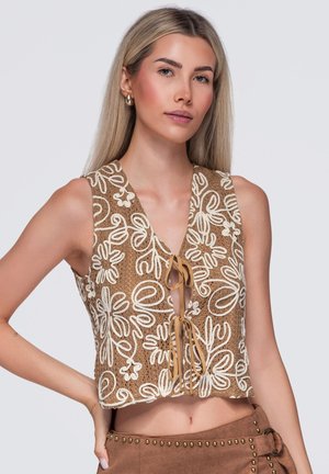 Giovane donna con lunghi capelli biondi che indossa un top marrone senza maniche con motivi floreali bianchi e nodo frontale, abbinato a pantaloni marroni con borchie.