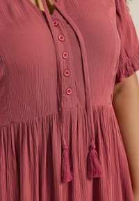 Ruusunpunainen mekko, jossa on teksturoitu kangas, viisi vaaleanpunaista nappia edessä ja solmimisnauha, jossa on tupsut kaula-aukossa.