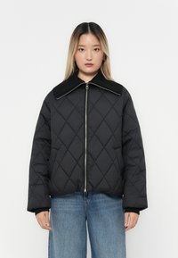 Giacca nera imbottita con un ampio collo a costine, chiusura lampo frontale e maniche lunghe. Indossata sopra jeans di denim chiaro.